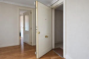 Puerta de entrada acorazada blanca abierta, con el canto dorado y la cerradura multipunto visibles